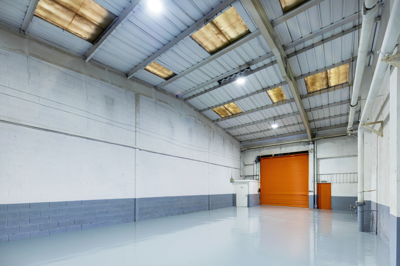 Large empty warehouse with white and grey painted walls, a polished floor, high ceiling, and an orange roller shutter door at the back.