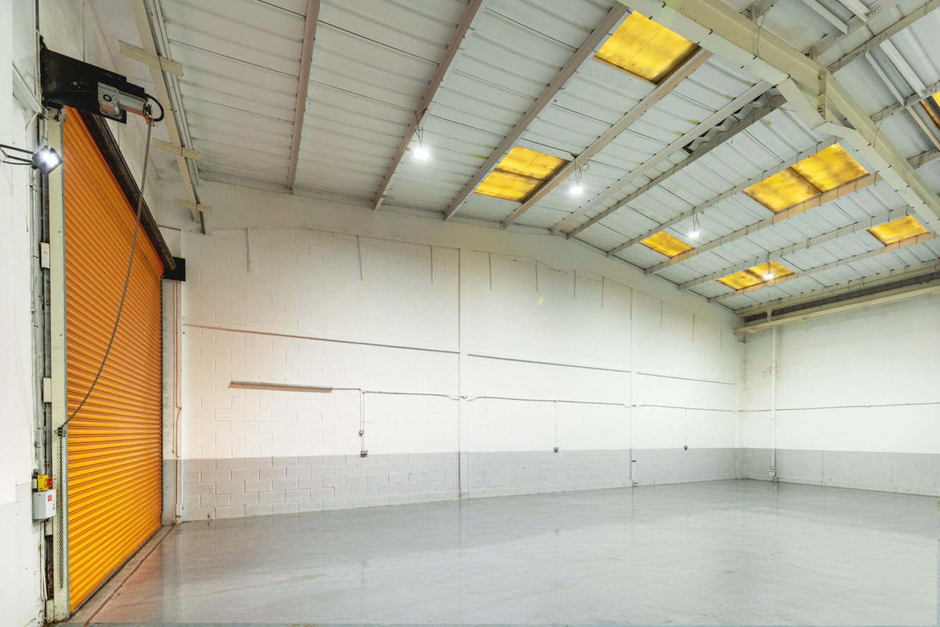 Empty industrial warehouse with white and grey walls, polished concrete floor, high ceiling, and an orange roller shutter door on the left. Bright ceiling lights and skylights are visible.