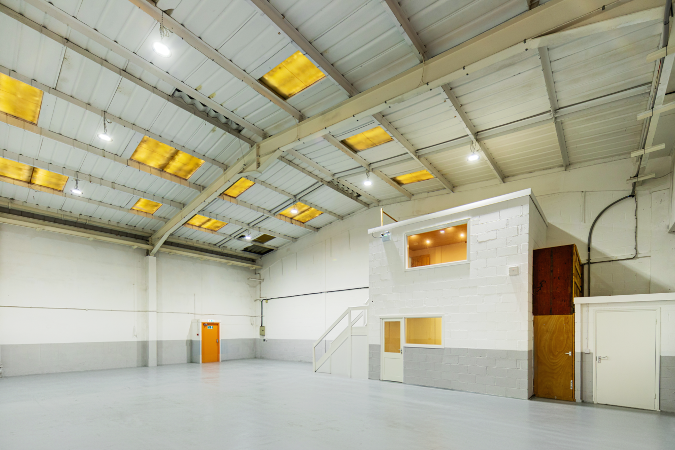 Spacious empty warehouse interior with high ceiling, concrete floors, white walls, a small two-storey office section, and a closed orange door.