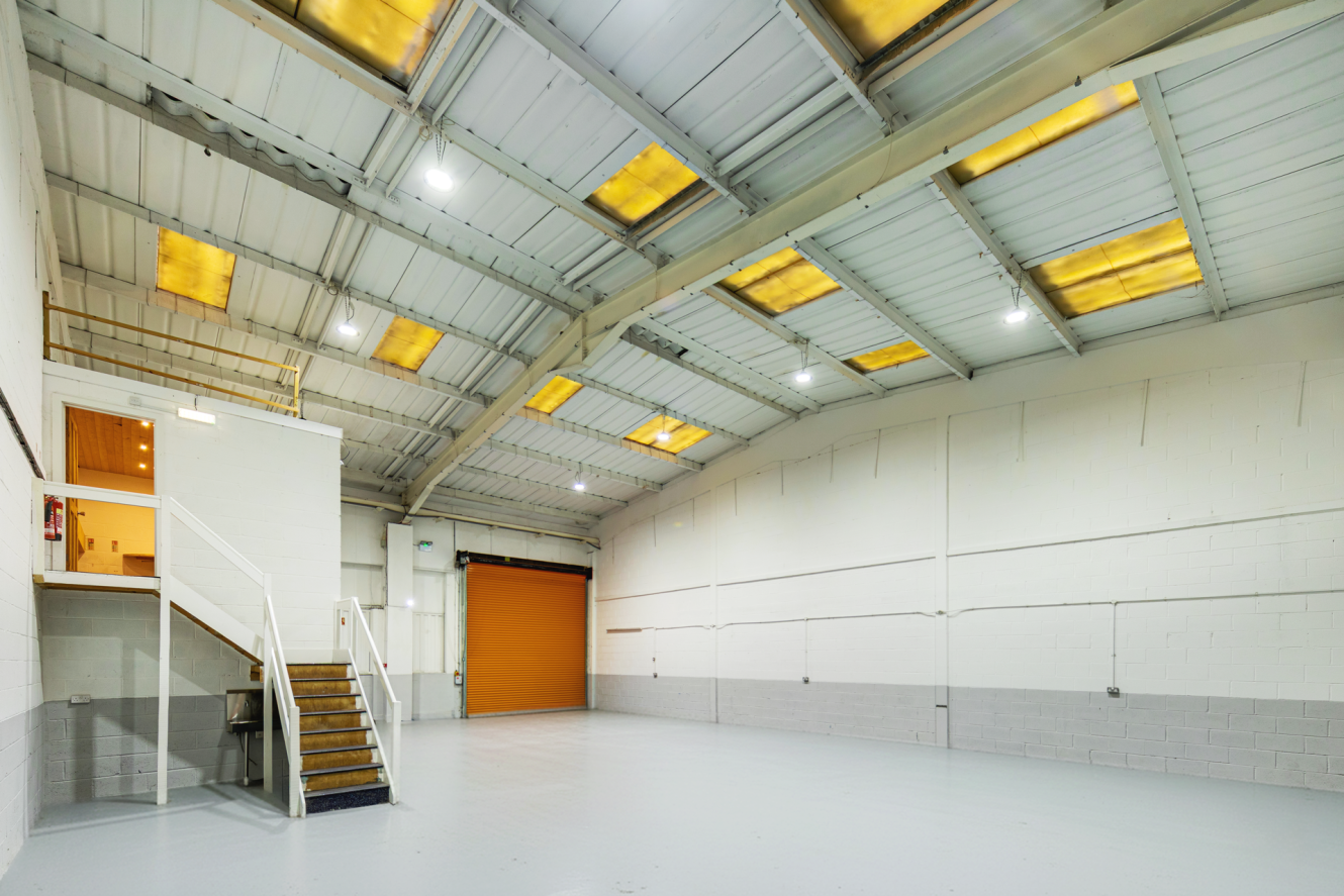 Empty industrial warehouse with white walls, high ceiling, overhead lights, a small office area with stairs, and a closed orange roller shutter door.