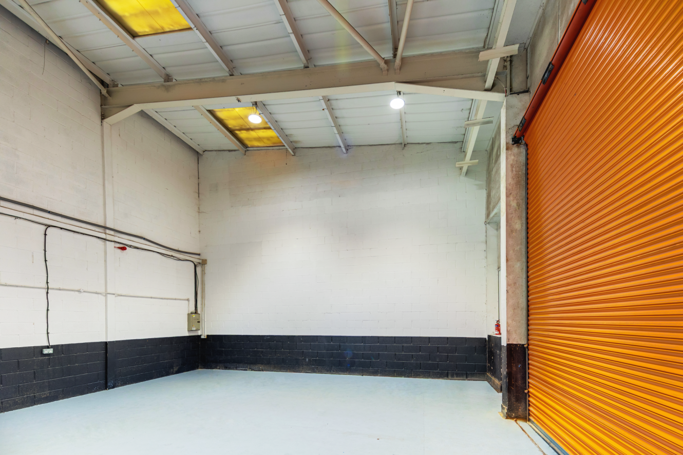 Empty warehouse interior with white walls, high ceiling, exposed beams, fluorescent lights, and an open orange roller shutter door on the right.