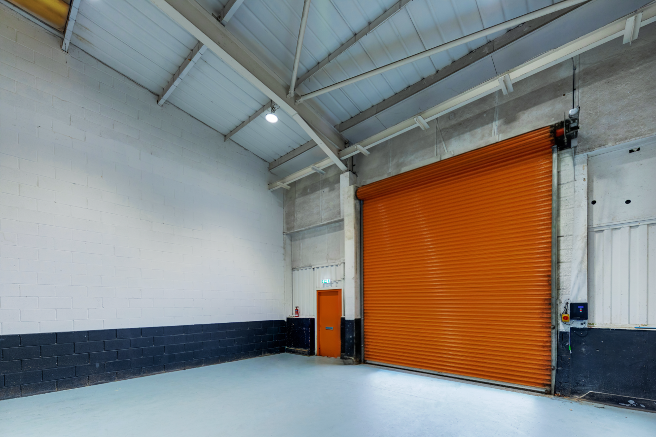 Interior of an empty warehouse with a high ceiling, white walls, a black lower wall trim, and a large closed orange roller shutter door next to a small orange entrance door.