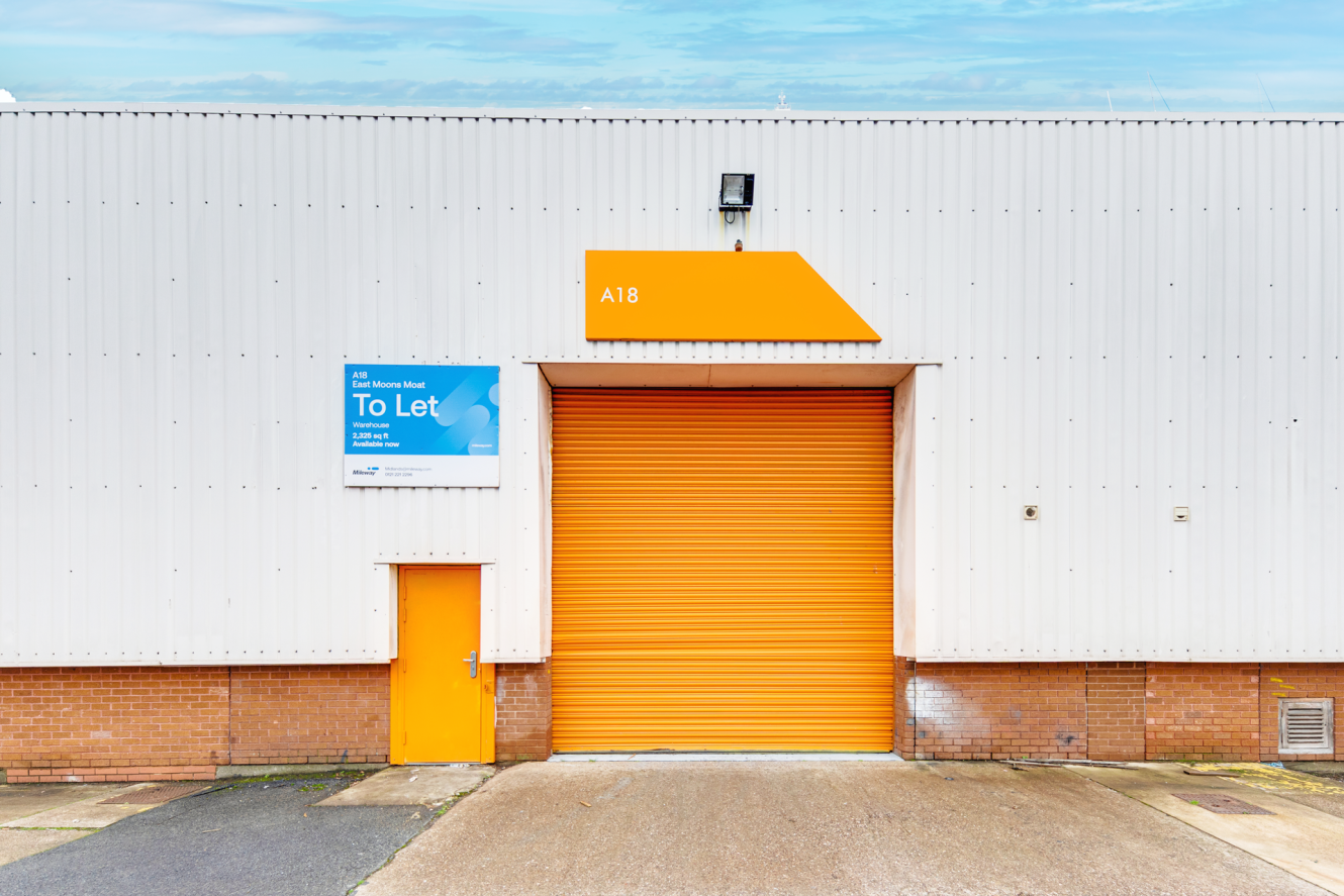 A large industrial unit with a white exterior, orange roller shutter door, and an orange side door; sign reads To Let with unit number A18 above the shutter.