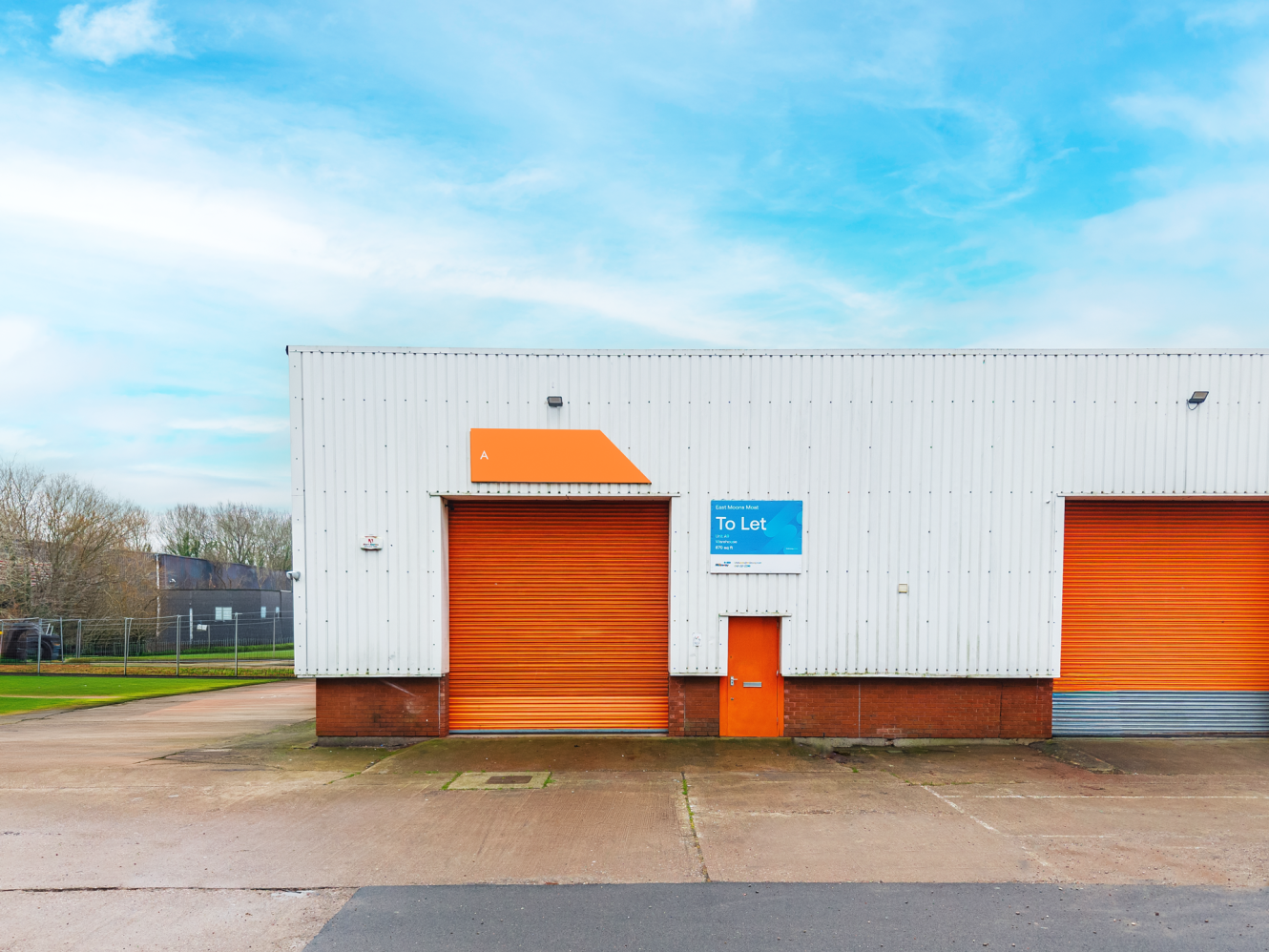 A warehouse unit with orange doors and a “To Let” sign on the front, set in an industrial area under a blue sky.