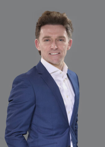 A man in a blue suit and white shirt stands against a plain grey background, smiling at the camera.