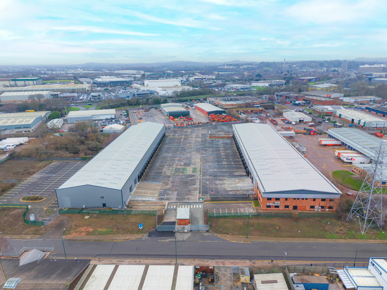 Aerial view of an industrial estate with large warehouses, open car parks, scattered vehicles, and surrounding commercial buildings under a cloudy sky.