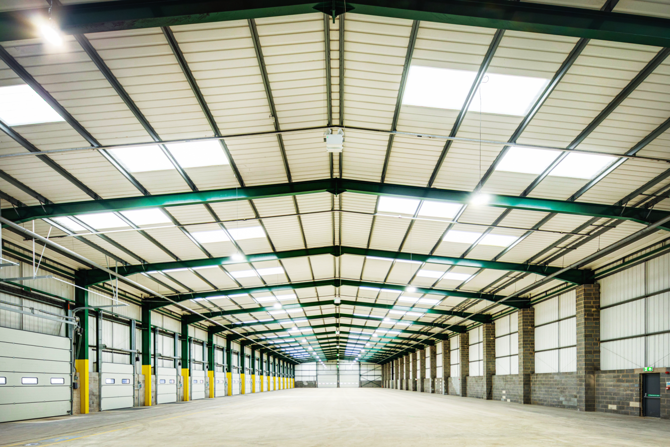Interior of a large, empty warehouse with high ceilings, skylights, overhead lights, and rows of closed loading bay doors on both sides.