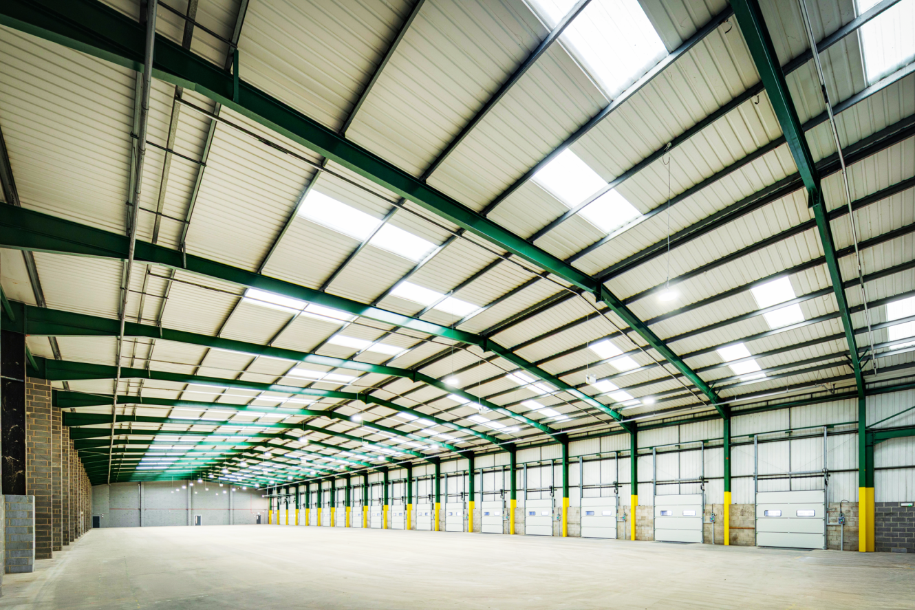 Interior of an empty, spacious warehouse with a high ceiling, skylights, and multiple closed loading bay doors along one side.