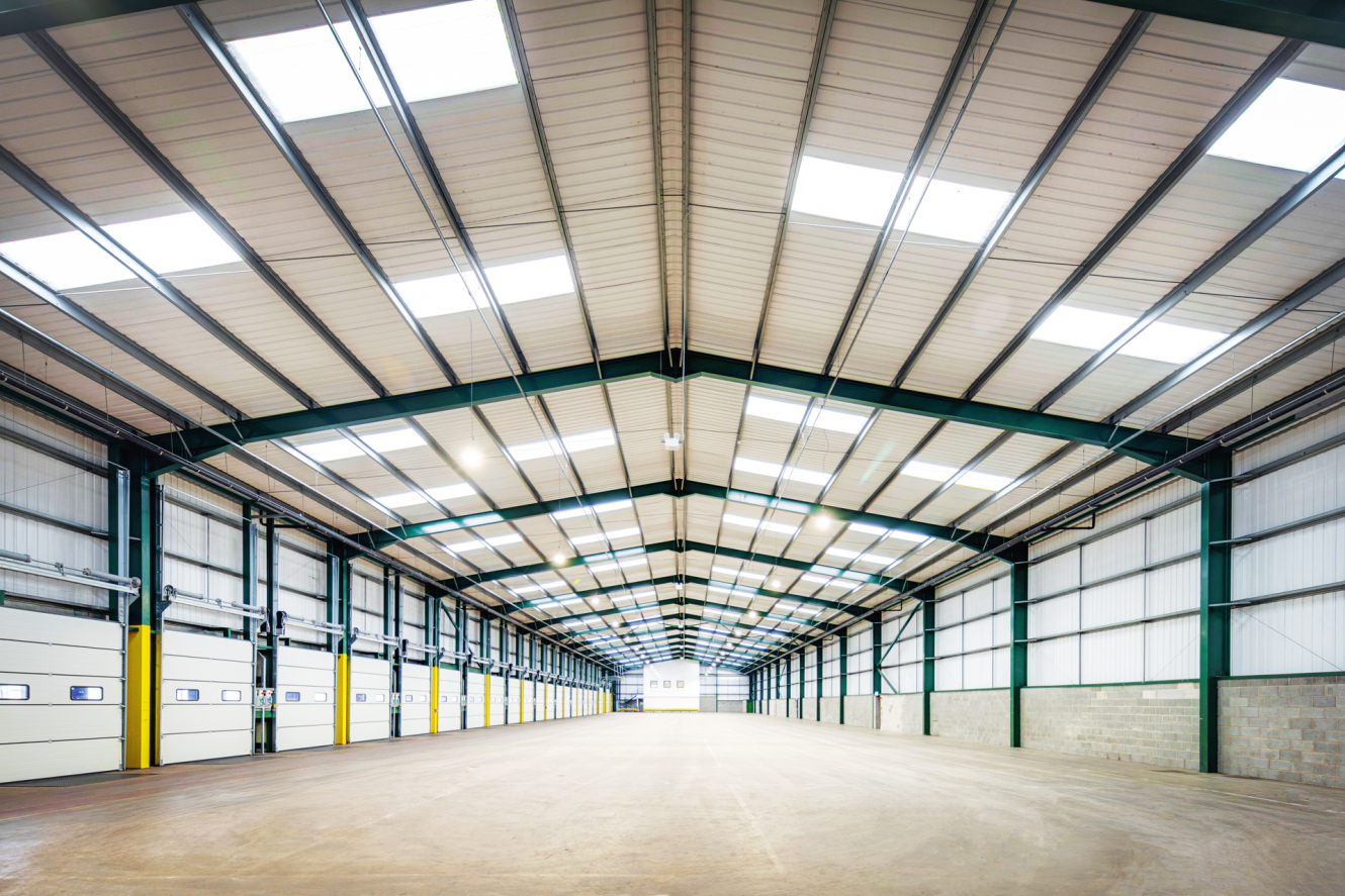 Empty, well-lit warehouse interior with a high metal ceiling, skylights, concrete floor, and large overhead doors along the left wall.