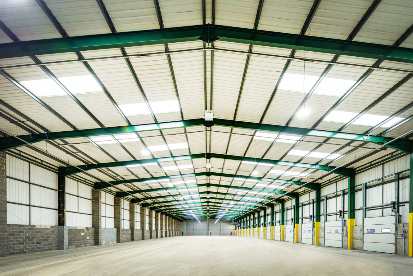 Wide, empty warehouse interior with high ceilings, metal beams, concrete floor, and multiple loading bay doors along one wall. Bright overhead lighting illuminates the space.