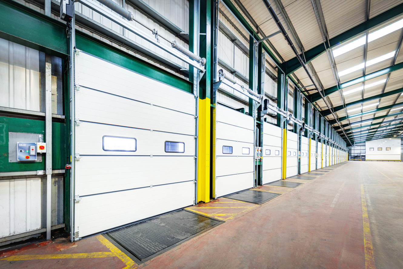 Interior view of a modern warehouse with multiple closed loading bay doors, green steel framing, and a clean, empty concrete floor.
