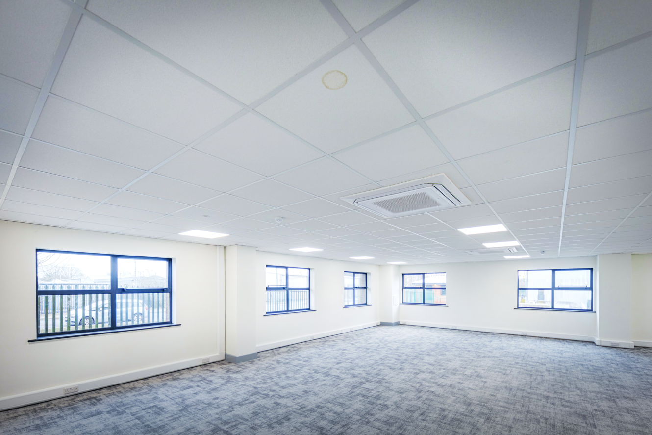 Empty office space with white walls, multiple windows, a grey carpeted floor, and a suspended ceiling with built-in lights and an air conditioning unit.