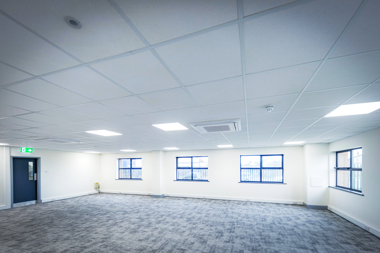 Empty office space with grey carpet, white walls, ceiling lights, air conditioning unit, and several windows letting in natural light.