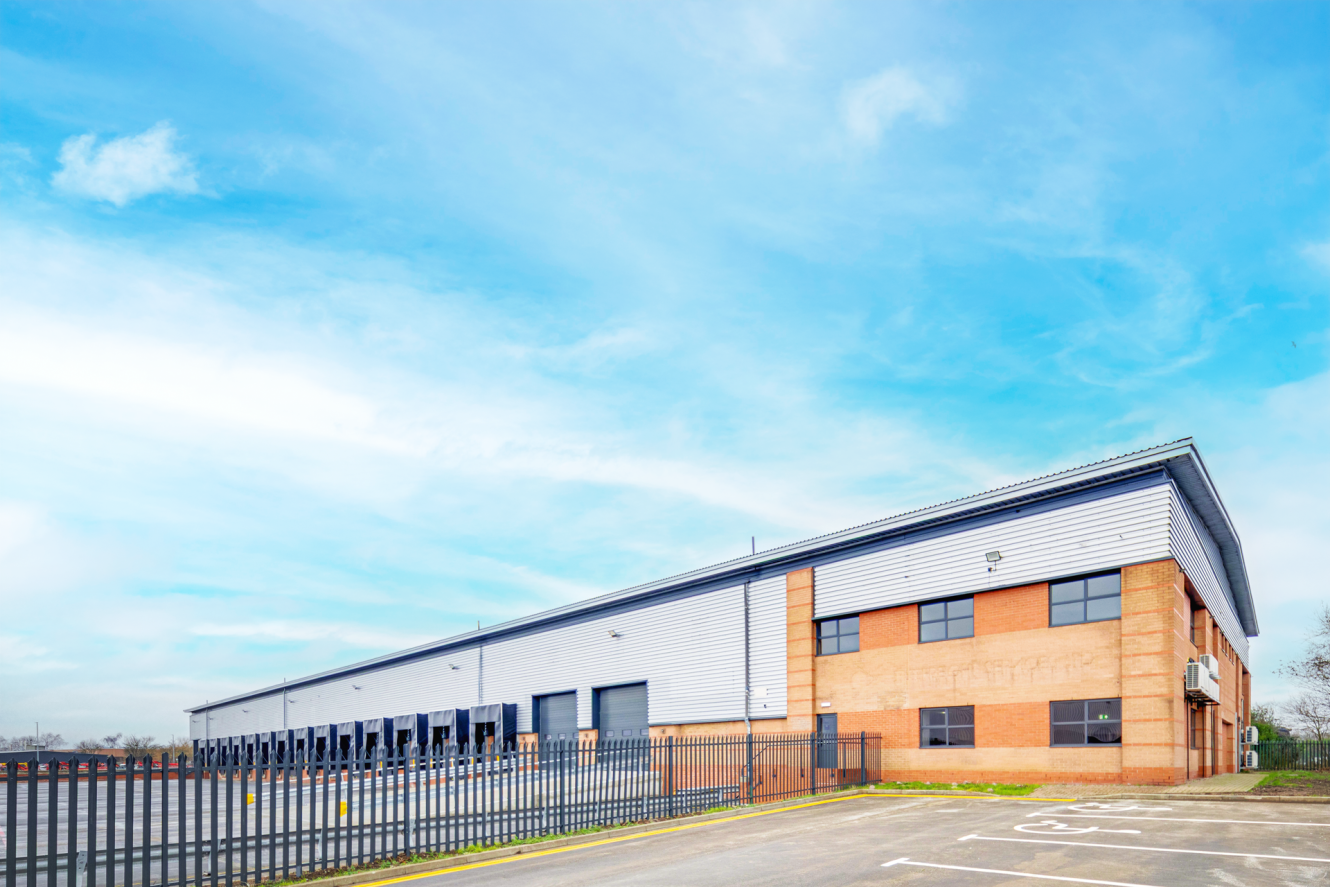 A large, modern industrial warehouse with a brick and metal facade, multiple loading bays, and a black metal fence in front, under a partly cloudy sky.