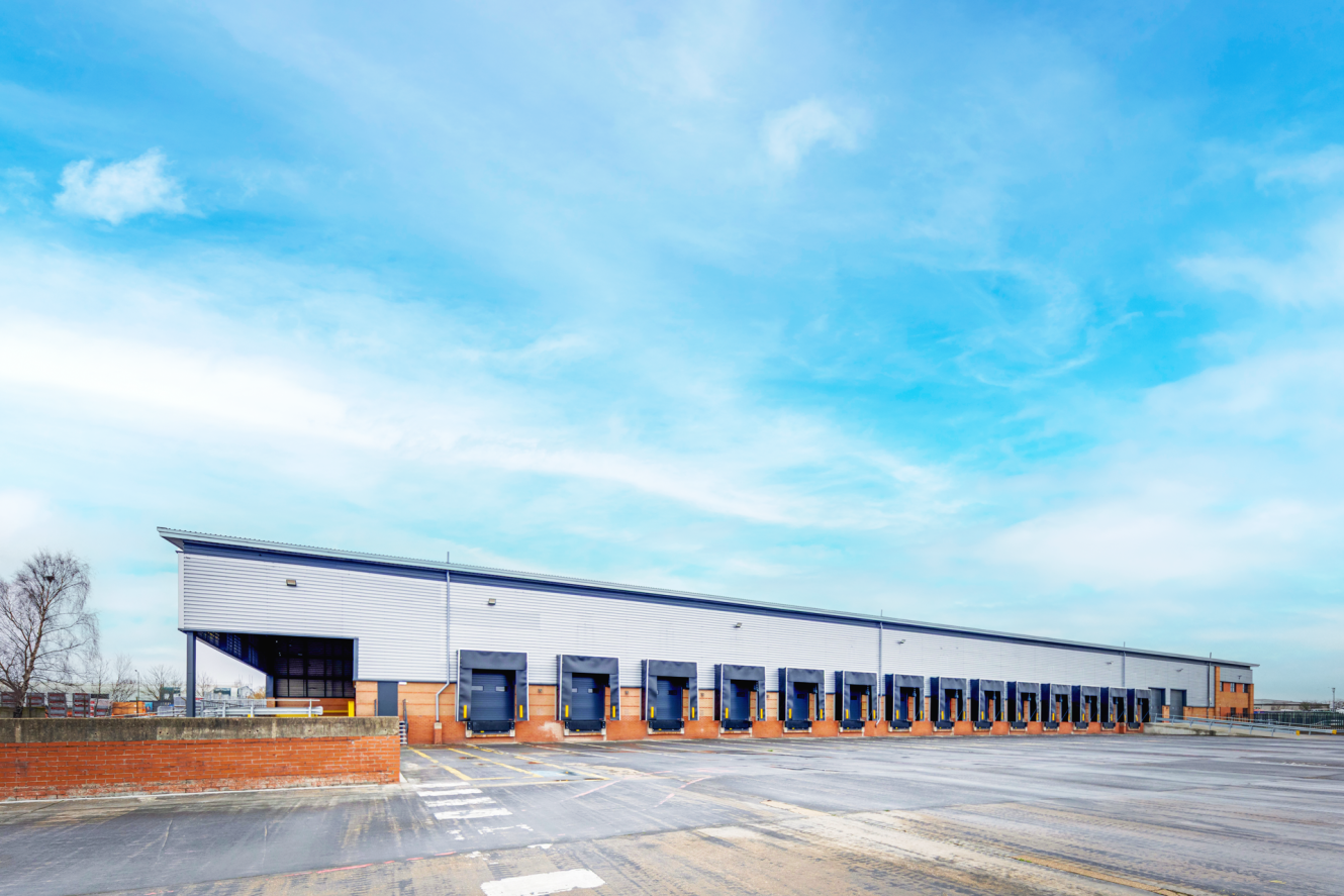 A large industrial warehouse with multiple loading bays and an empty tarmacked yard under a blue sky.