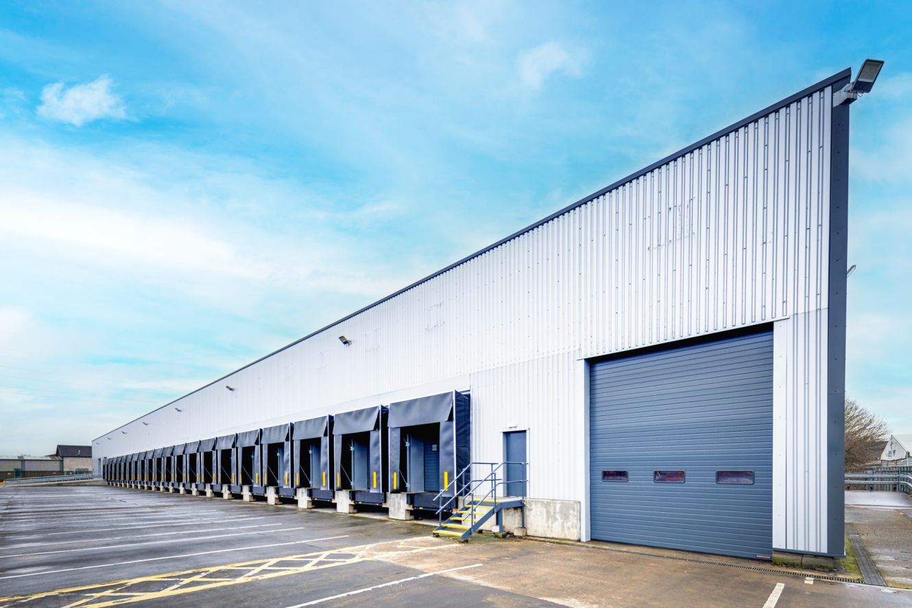 Large industrial warehouse with multiple loading bays and a closed roller shutter door, set beside an empty car park under a clear blue sky.