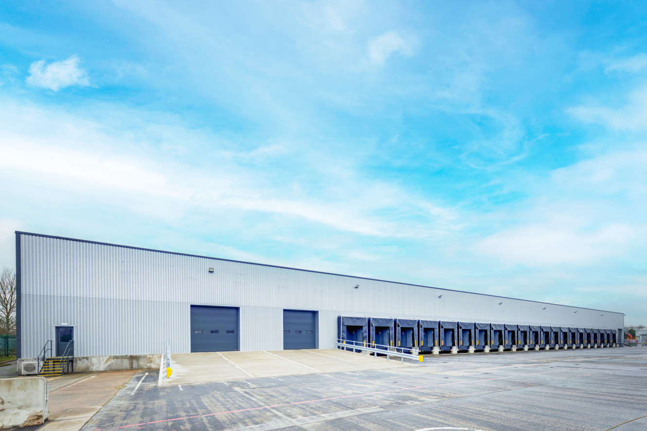 Large warehouse building with multiple loading bay doors and a wide concrete car park under a partly cloudy sky.