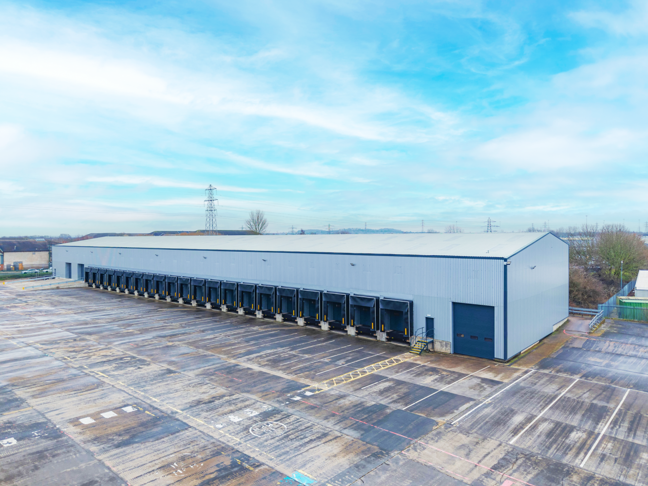 A large industrial warehouse with multiple loading bays and an empty tarmac car park in front, under a partly cloudy sky.
