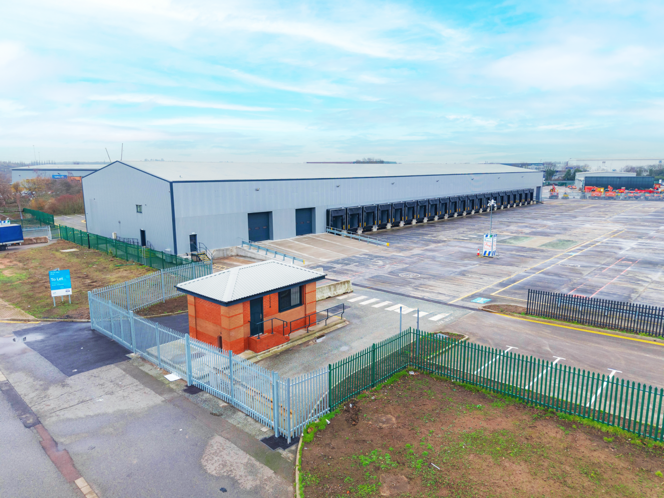 A large industrial warehouse with multiple loading bays, a small brick security building, and fenced perimeter under a partly cloudy sky.