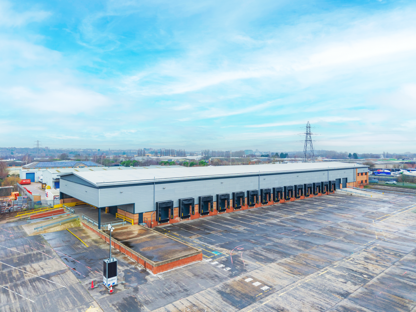 A large industrial warehouse with multiple loading bays is surrounded by an empty paved car park under a partly cloudy sky.