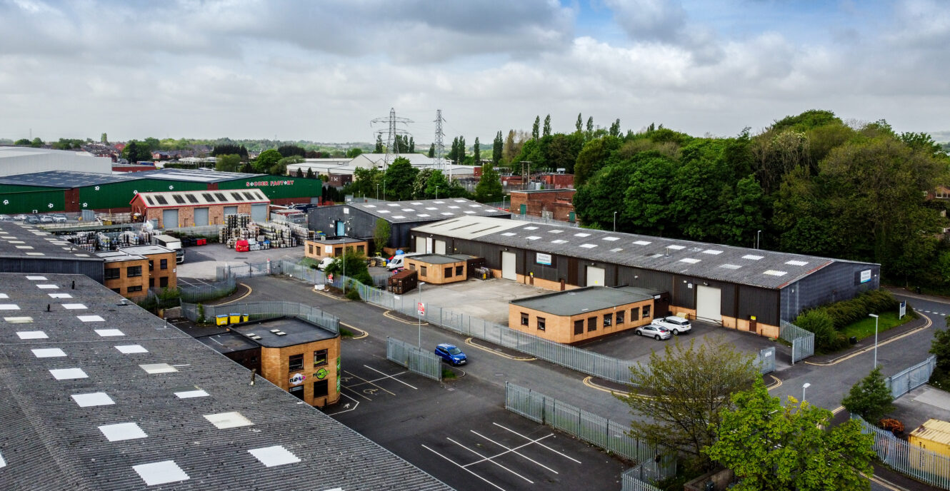 Aerial view of an industrial estate with warehouses, office buildings, parked vehicles, and surrounding trees under a partly cloudy sky.