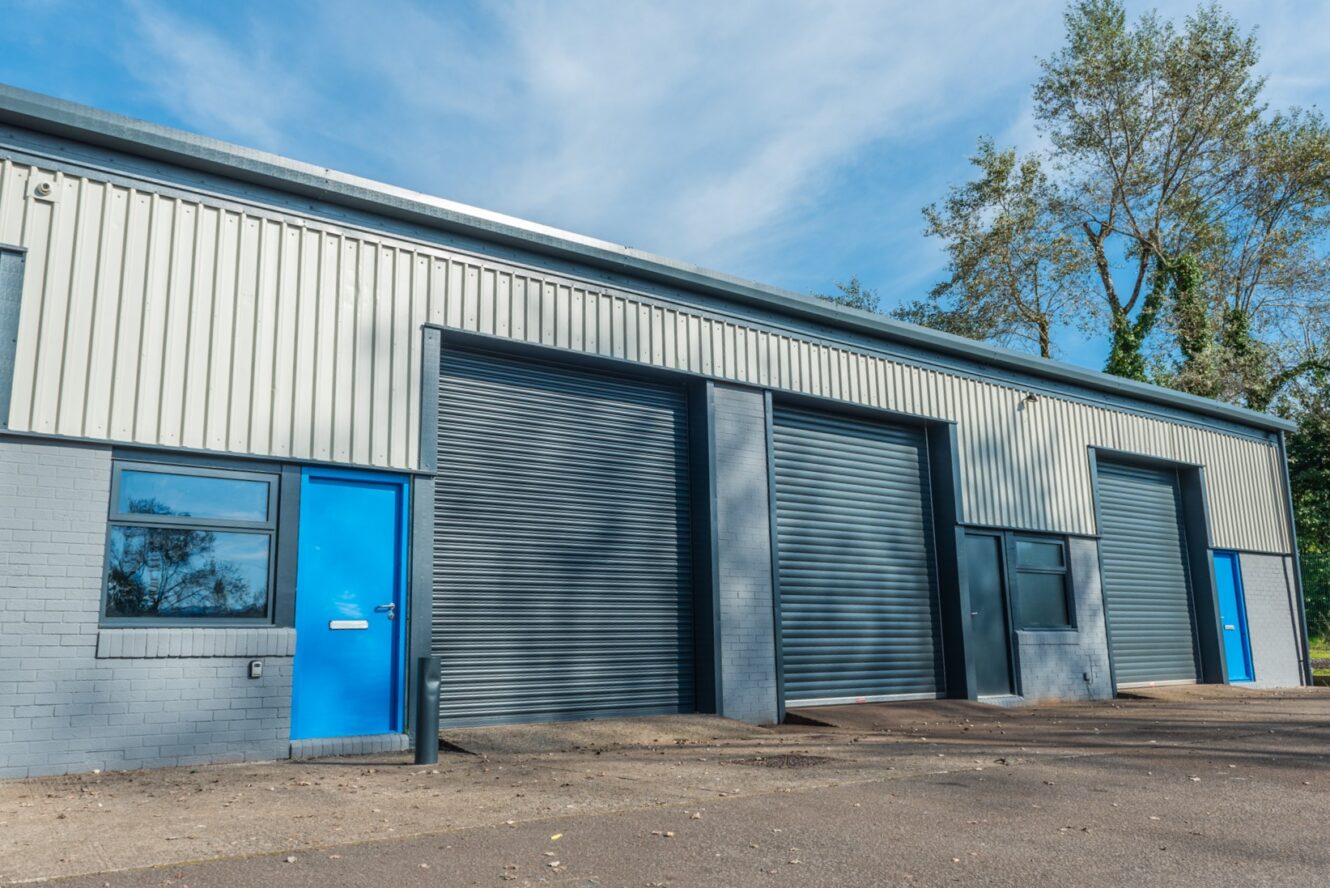 A row of industrial units with blue doors and grey roller shutters, situated in a paved area with trees visible in the background under a partly cloudy sky.