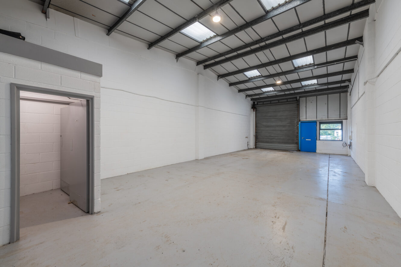 Empty industrial warehouse with white brick walls, high ceiling, overhead lights, a metal shutter door, blue entrance door, and a small separate room to the left.