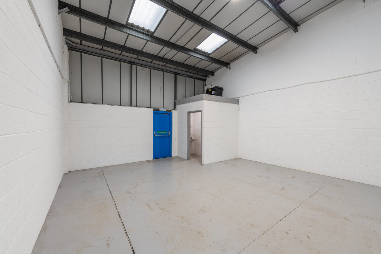 Empty industrial room with white brick walls, grey concrete floor, exposed metal ceiling, a blue door, and a small interior room with an open door. Fluorescent lights are on the ceiling.