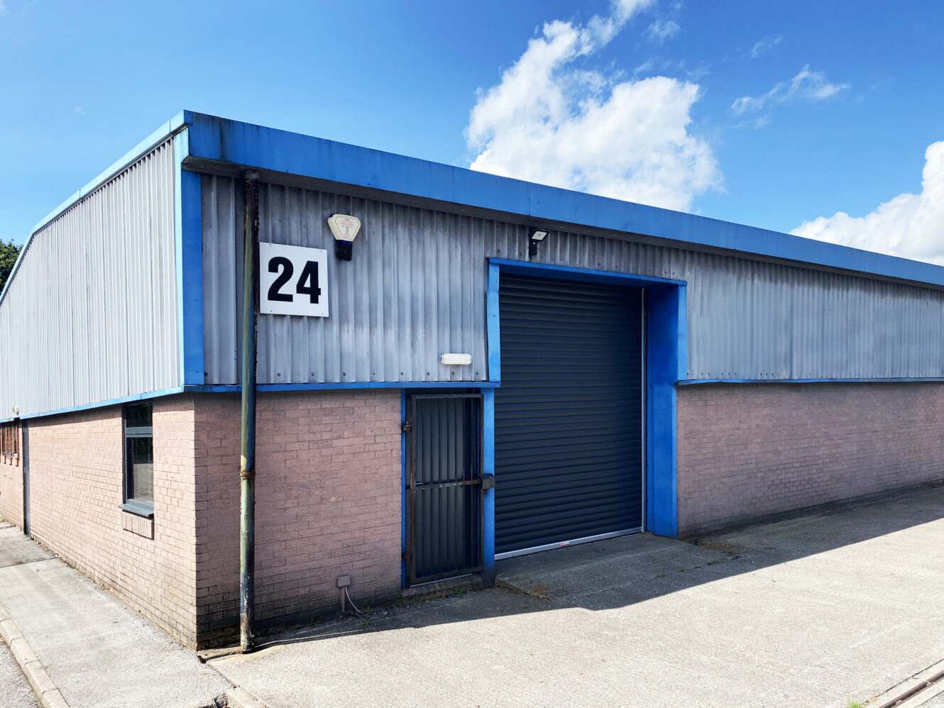A light industrial warehouse with grey metal cladding, a blue trim, and a large roller shutter door. A sign with the number 24 is mounted beside a smaller pedestrian door.