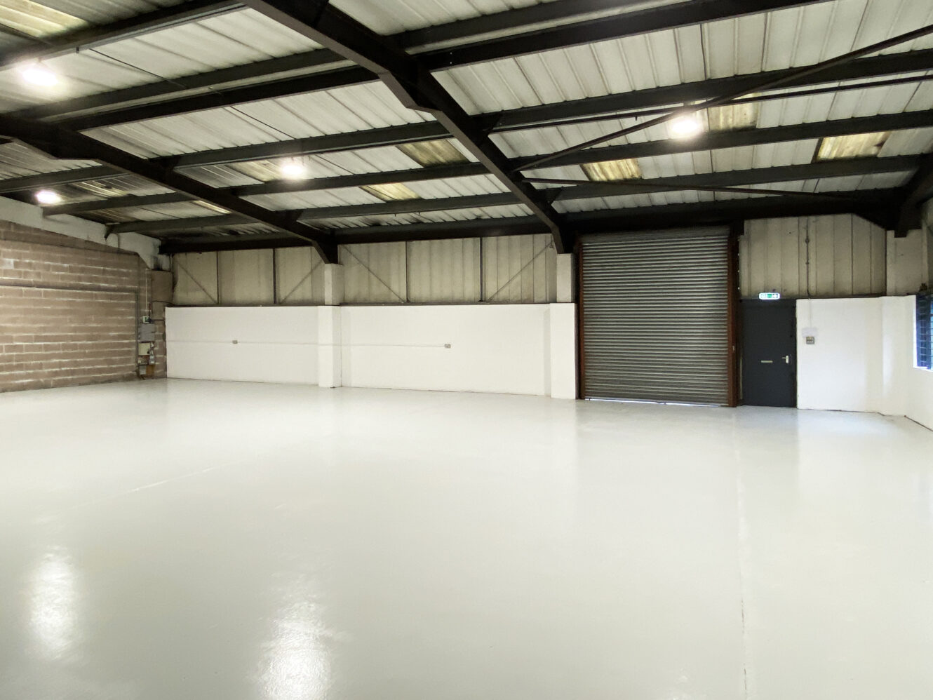 Empty industrial warehouse interior with polished concrete floor, high ceiling, exposed beams, brick and white walls, a large roller shutter door, and an emergency exit door.