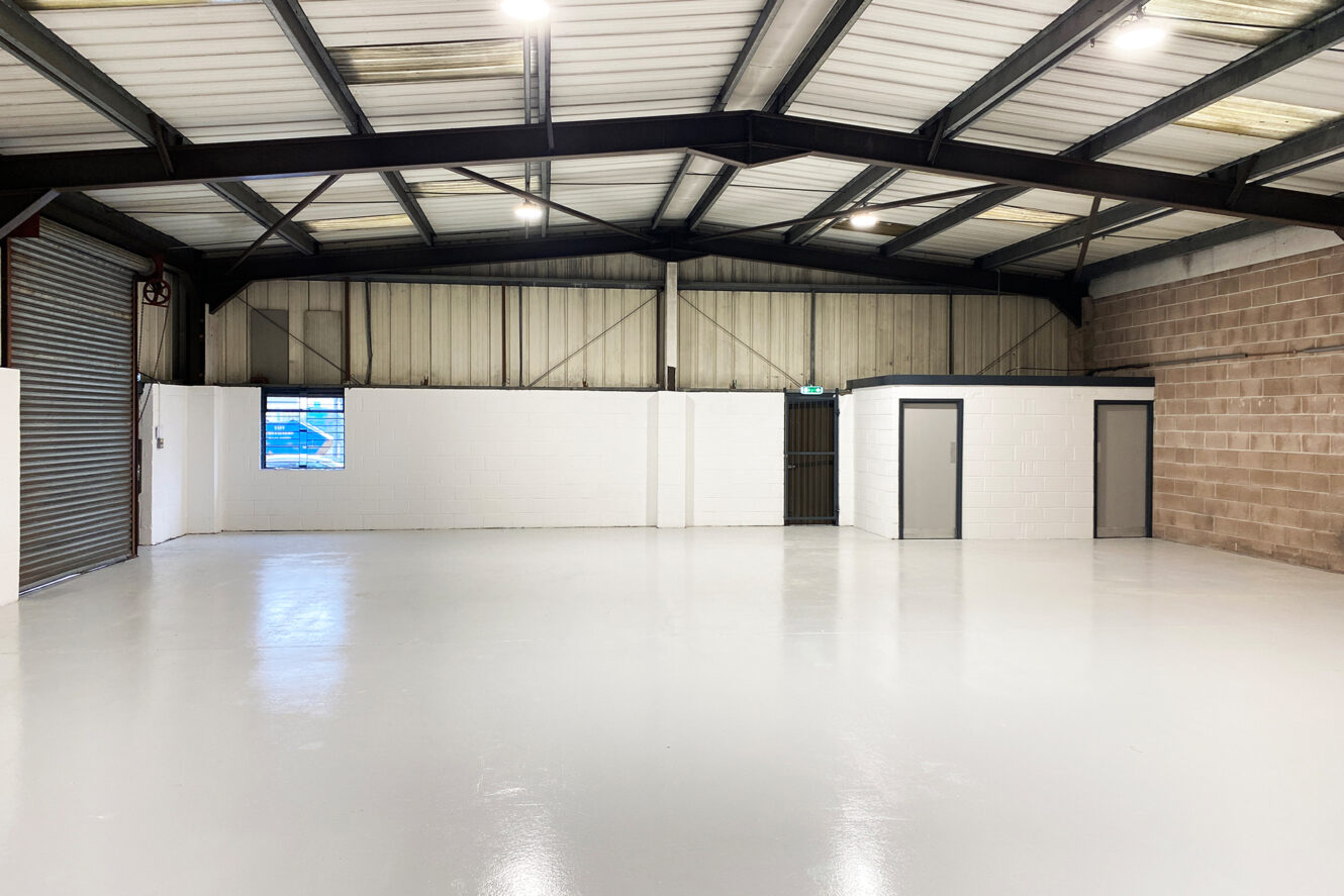 Interior of an empty industrial warehouse with a polished floor, metal roof beams, a large roller shutter, and two small rooms with doors inside.