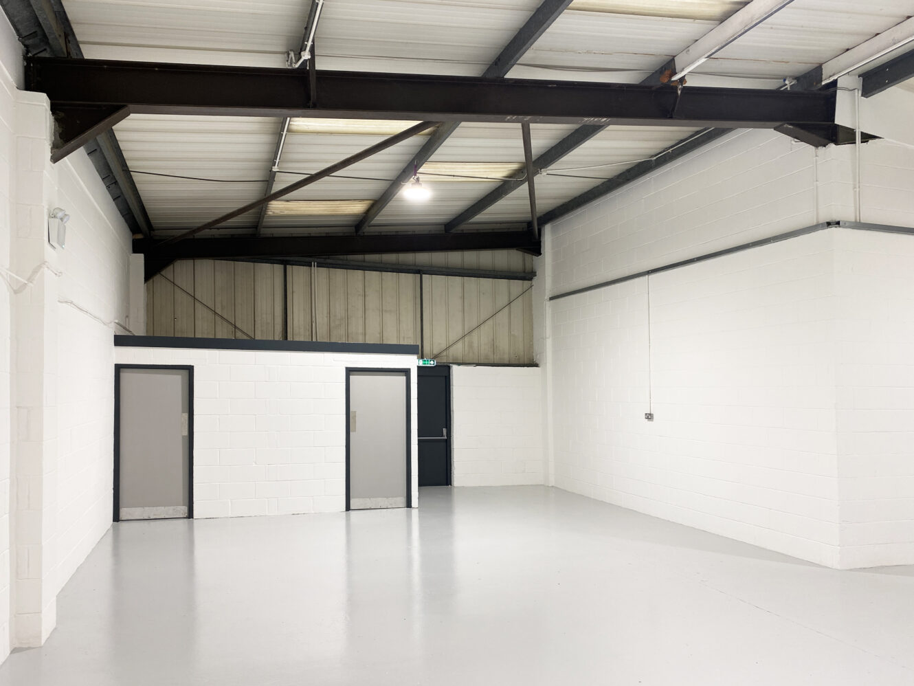 Empty industrial warehouse space with white walls, grey doors, exposed ceiling beams, and a concrete floor. Overhead lighting illuminates the area.