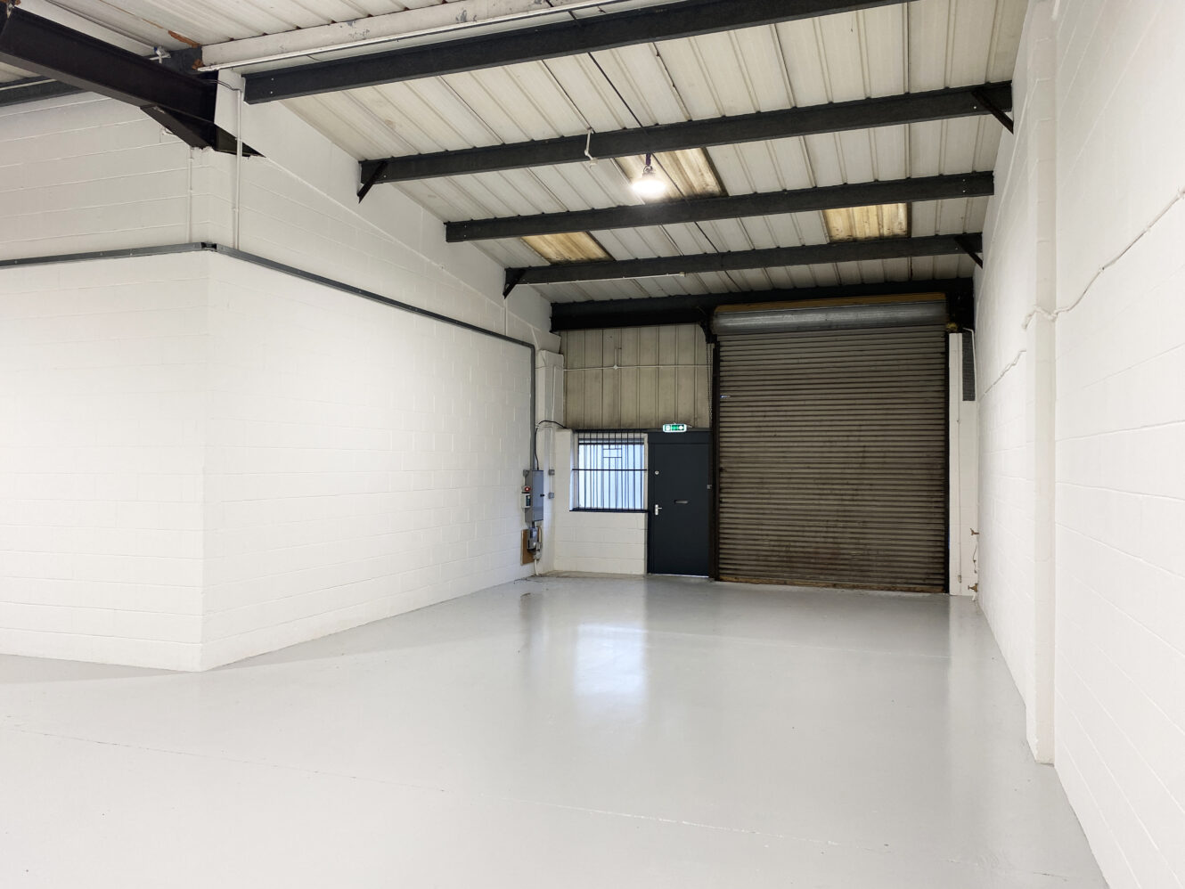 Empty industrial warehouse space with white walls, a grey metal roller shutter door, a single door with a window, and exposed ceiling beams.