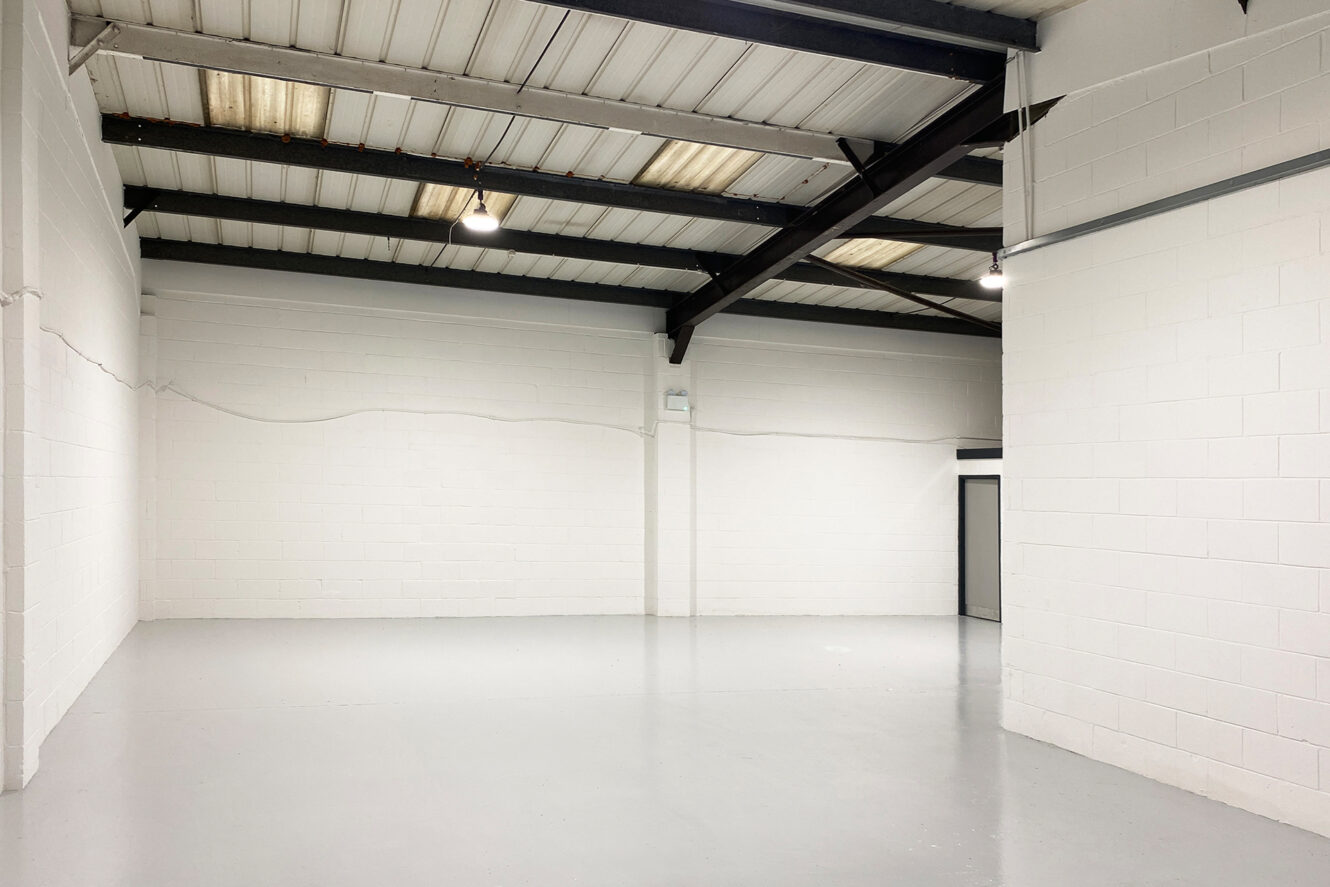 Empty industrial warehouse with white painted brick walls, exposed steel beams, fluorescent lighting, and a smooth grey floor.
