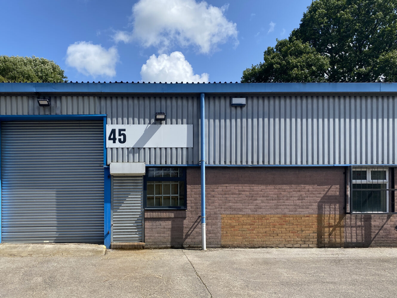 Industrial building with a large grey roller door, two windows, and the number 45 displayed above a small entrance; blue trim and trees in the background.