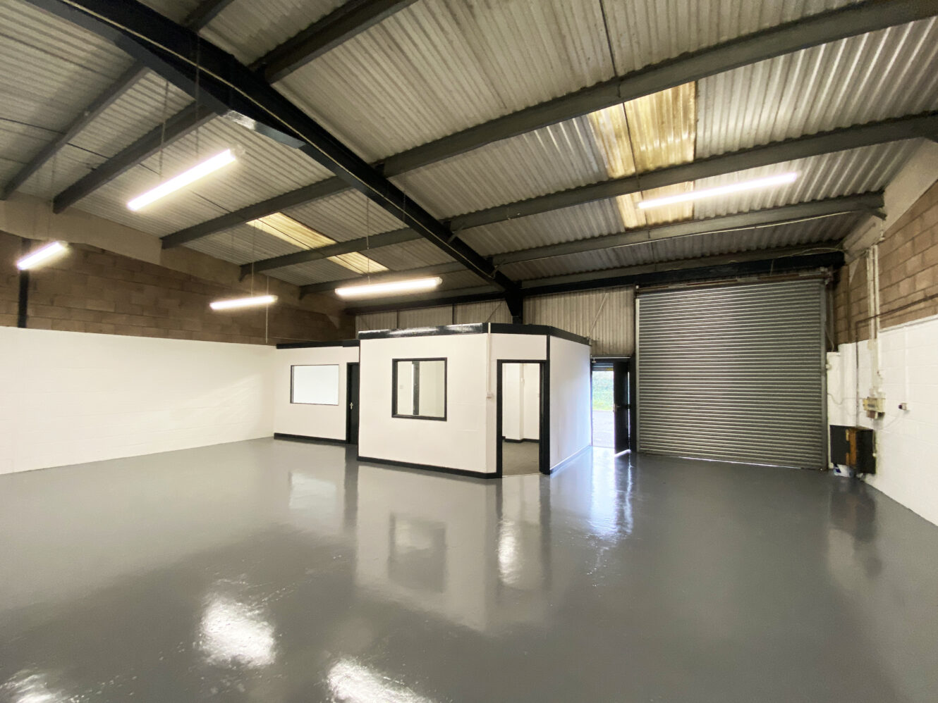 Interior of an empty industrial warehouse with a small office unit, polished grey floor, high ceiling, fluorescent lighting, and a large roller shutter door.