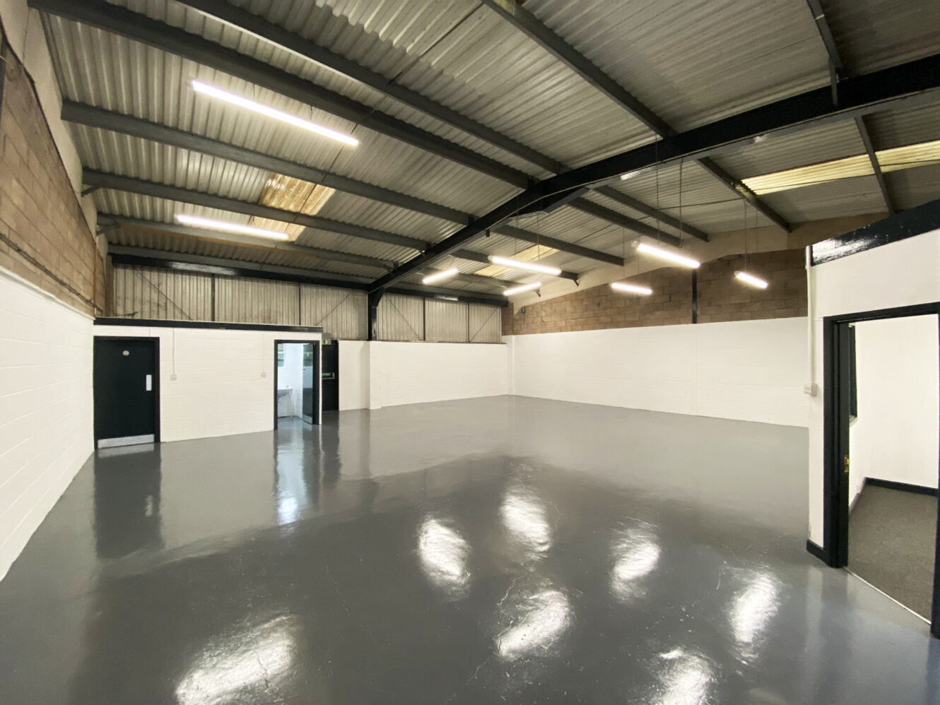 Spacious, empty industrial warehouse with polished grey concrete floor, exposed metal beams, brick and white walls, fluorescent ceiling lights, and two open doorways.