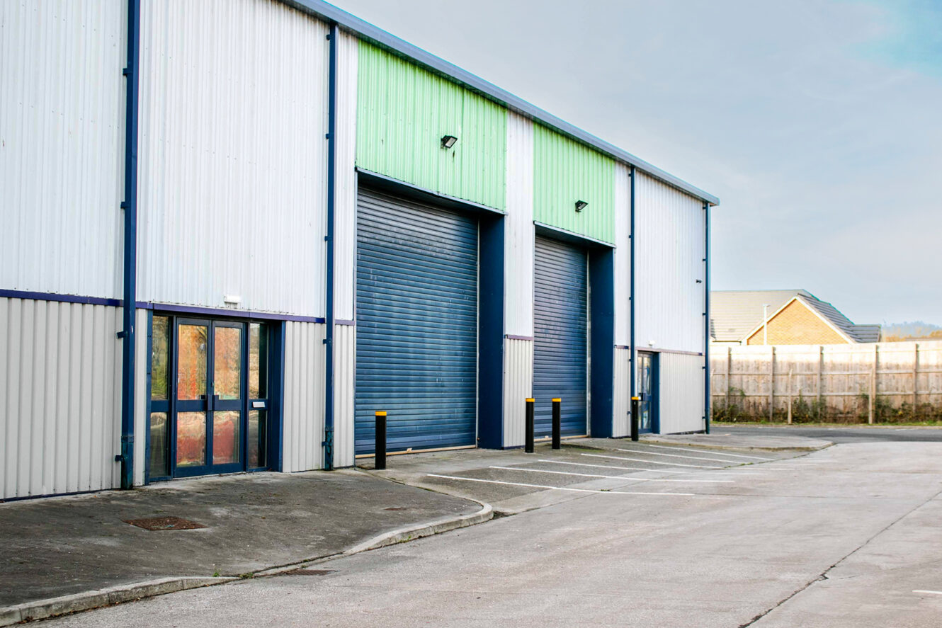 Exterior of a modern industrial warehouse with two large blue roller shutter doors and a glass entrance door, set beside an empty car park.