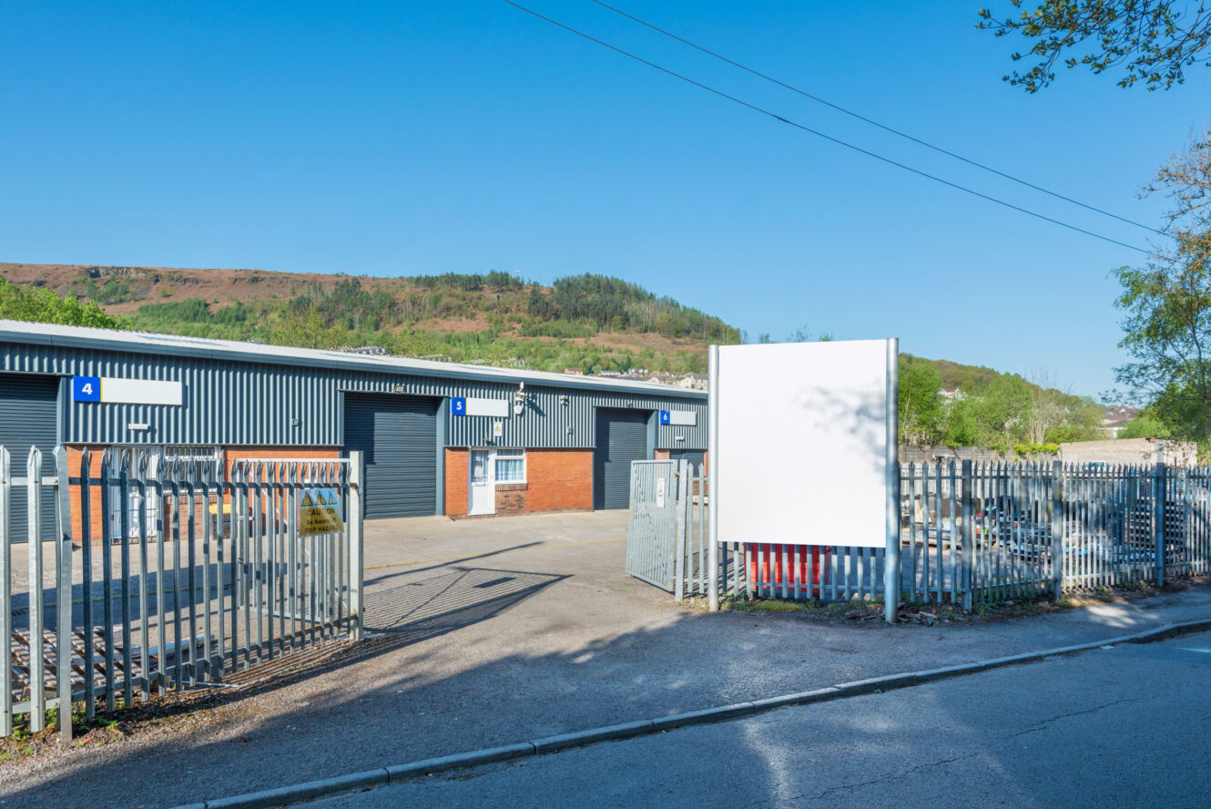 Industrial units with blue roller shutters and a blank white sign, surrounded by a metal fence on a sunny day with hills in the background.
