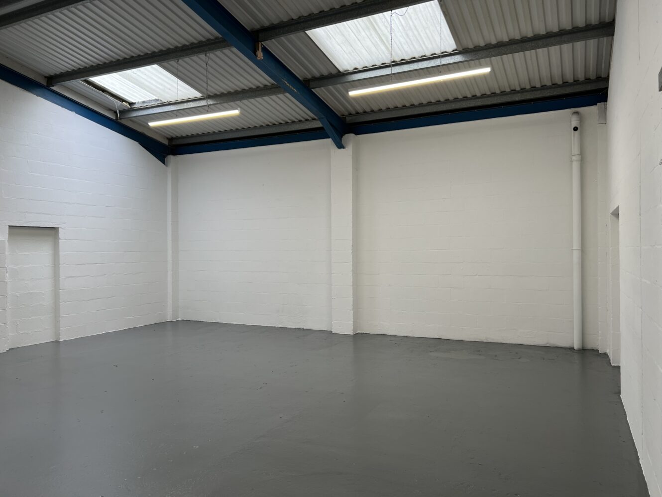 Empty industrial warehouse unit with white painted brick walls, grey painted concrete floor, skylights, and exposed blue ceiling beams.