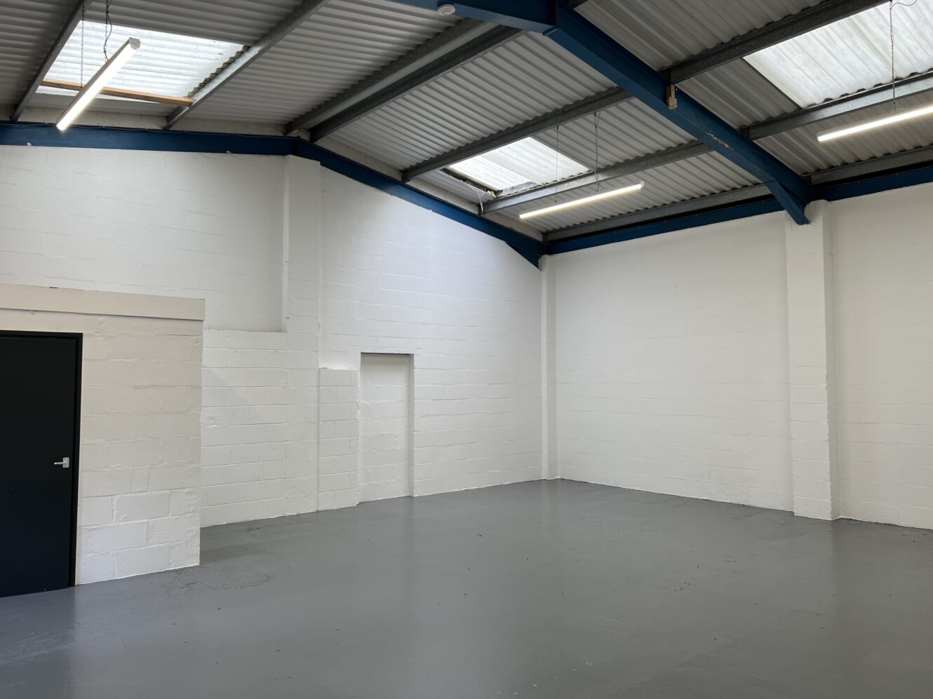 Empty industrial unit with white painted brick walls, grey concrete floor, dark door, high ceiling with blue beams, and skylights letting in natural light.