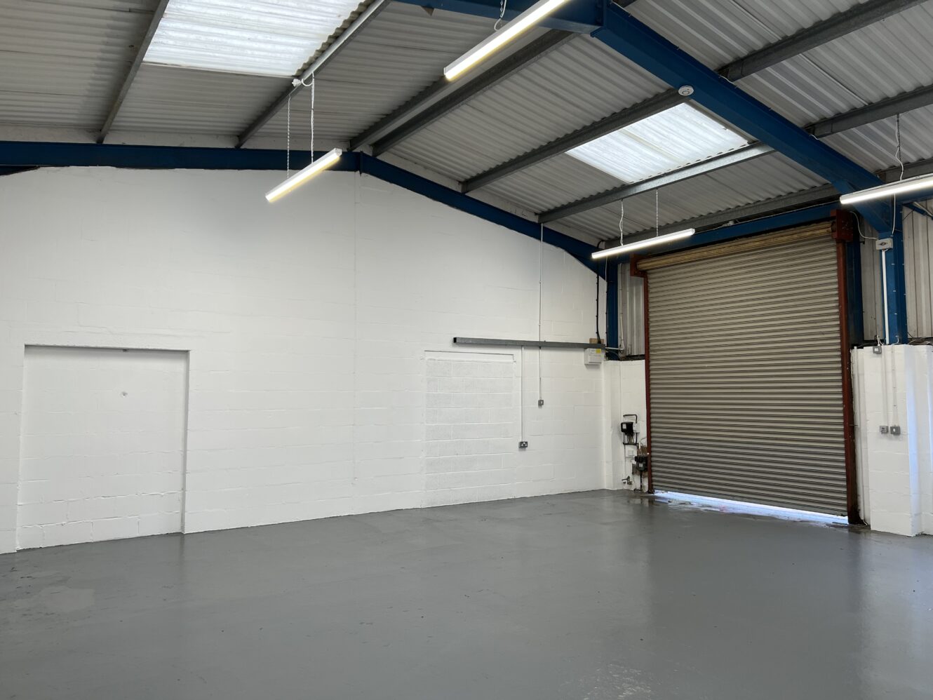 An empty industrial warehouse with white walls, grey flooring, exposed beams, fluorescent lights, and a partially open roller shutter door.