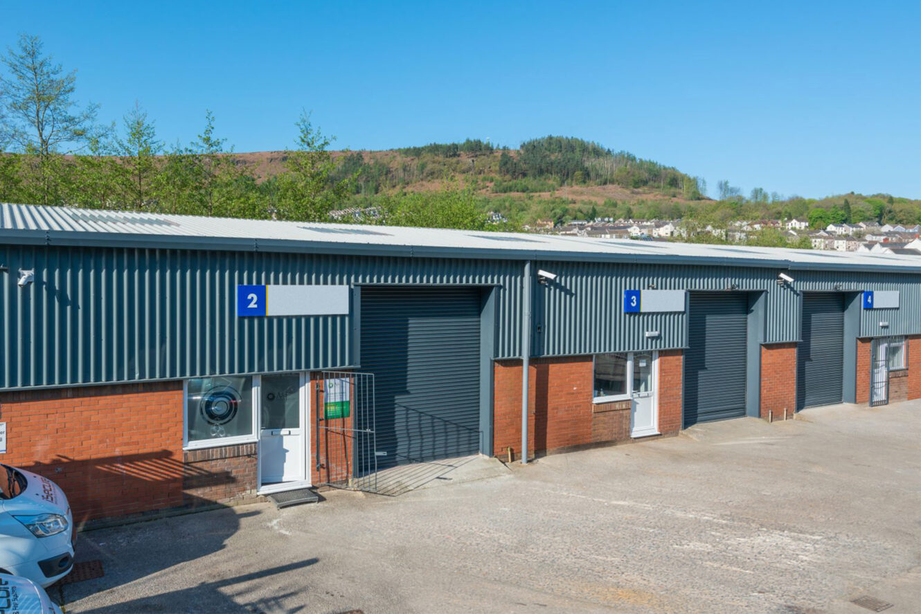 A row of industrial units with blue metal panels and brick lower walls, numbered 2 and 3, under a clear sky with a hill and trees in the background.