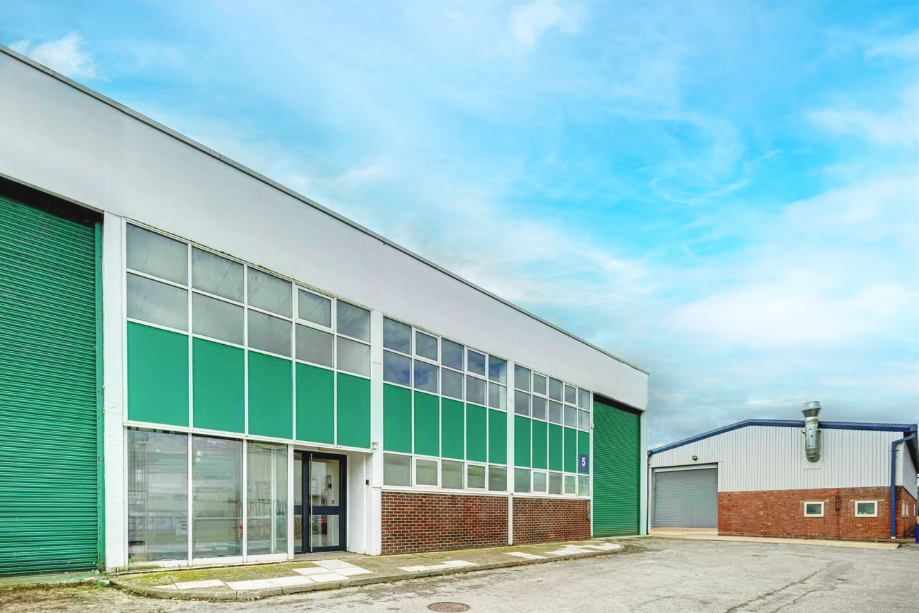 A large industrial warehouse with green and white exterior panels, glass windows, and a closed green roller shutter under a partly cloudy sky.
