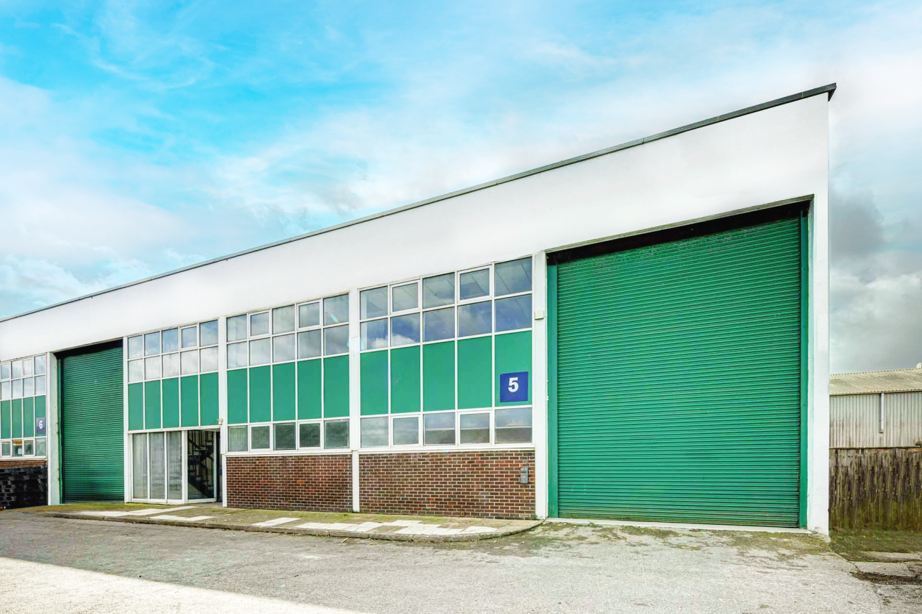 Industrial warehouse with green metal roller shutters and large windows, marked with the number 5, under a partly cloudy sky.