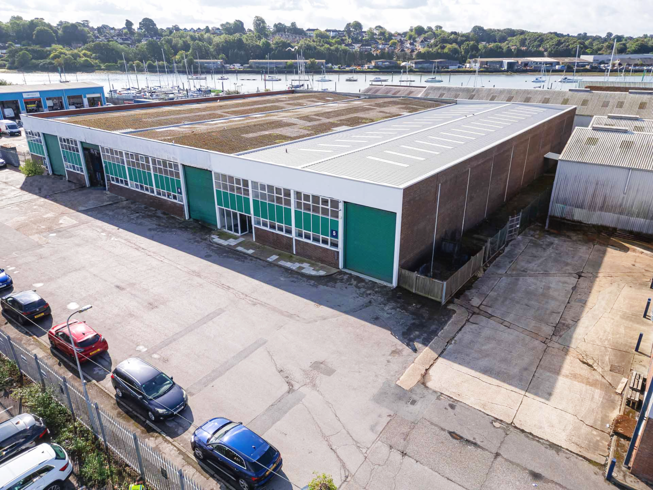 Aerial view of a large industrial warehouse with green doors, adjacent car park, and nearby river in the background.