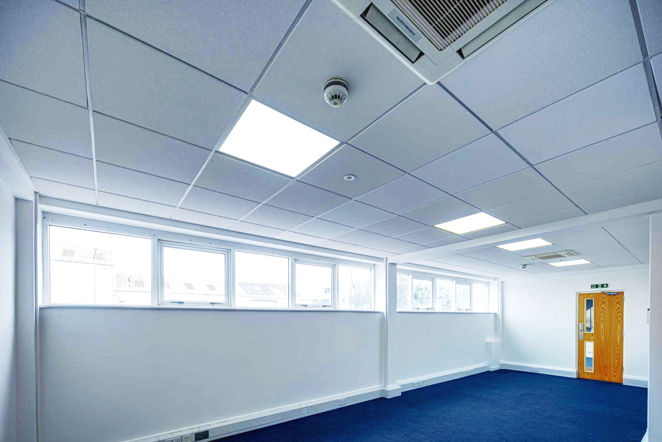 An empty office room with blue carpet, white walls, large windows, ceiling lights, and an air conditioning unit on the ceiling.