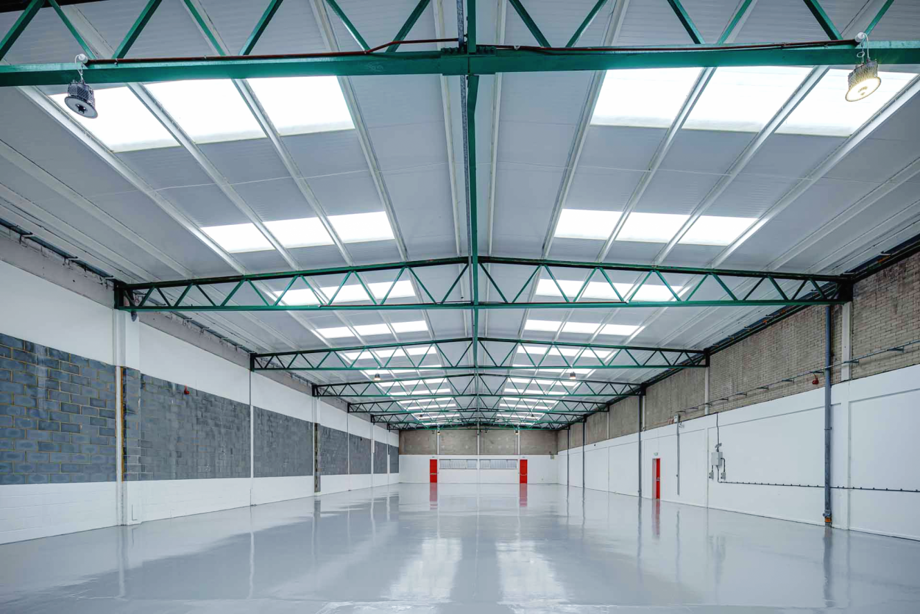 Empty industrial warehouse with polished concrete floor, skylights, exposed steel beams, and white walls with some brick and breeze block sections.