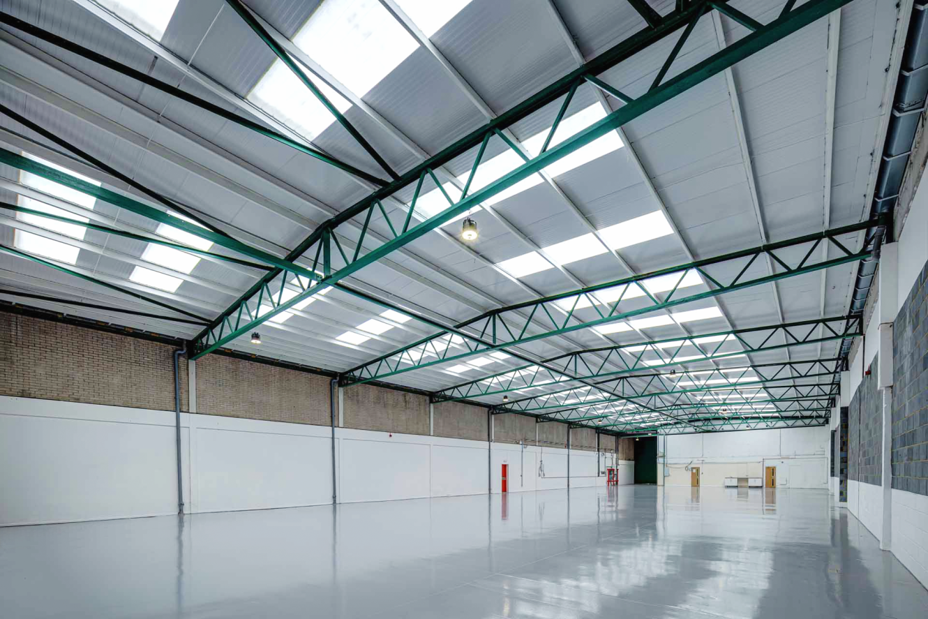 Interior of an empty industrial warehouse with high ceilings, exposed beams, skylights, and polished concrete floors. Walls are white with a few doors and fire safety equipment visible.