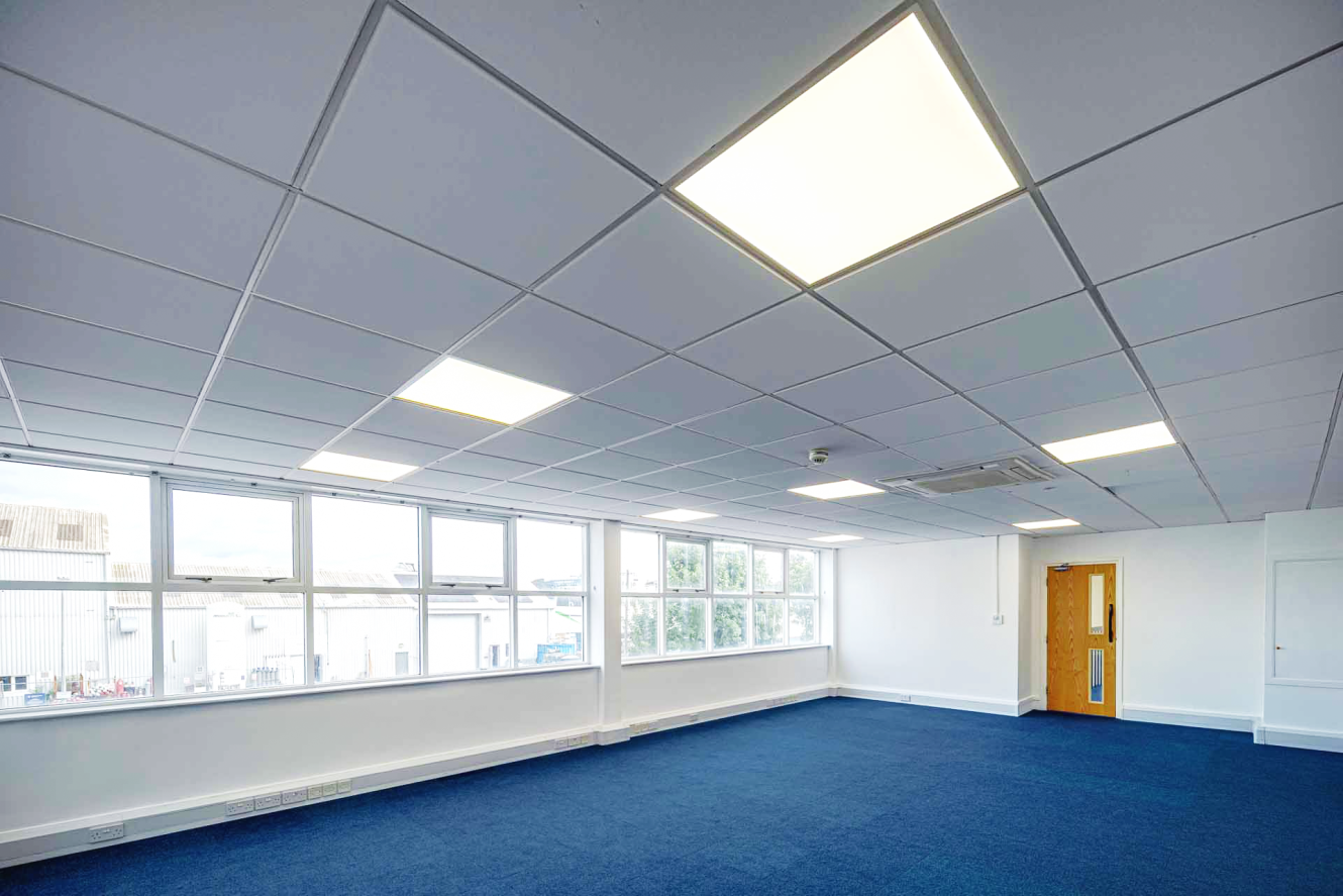 A spacious, empty office room with blue carpet, white walls, large windows, ceiling lights, and a closed wooden door.